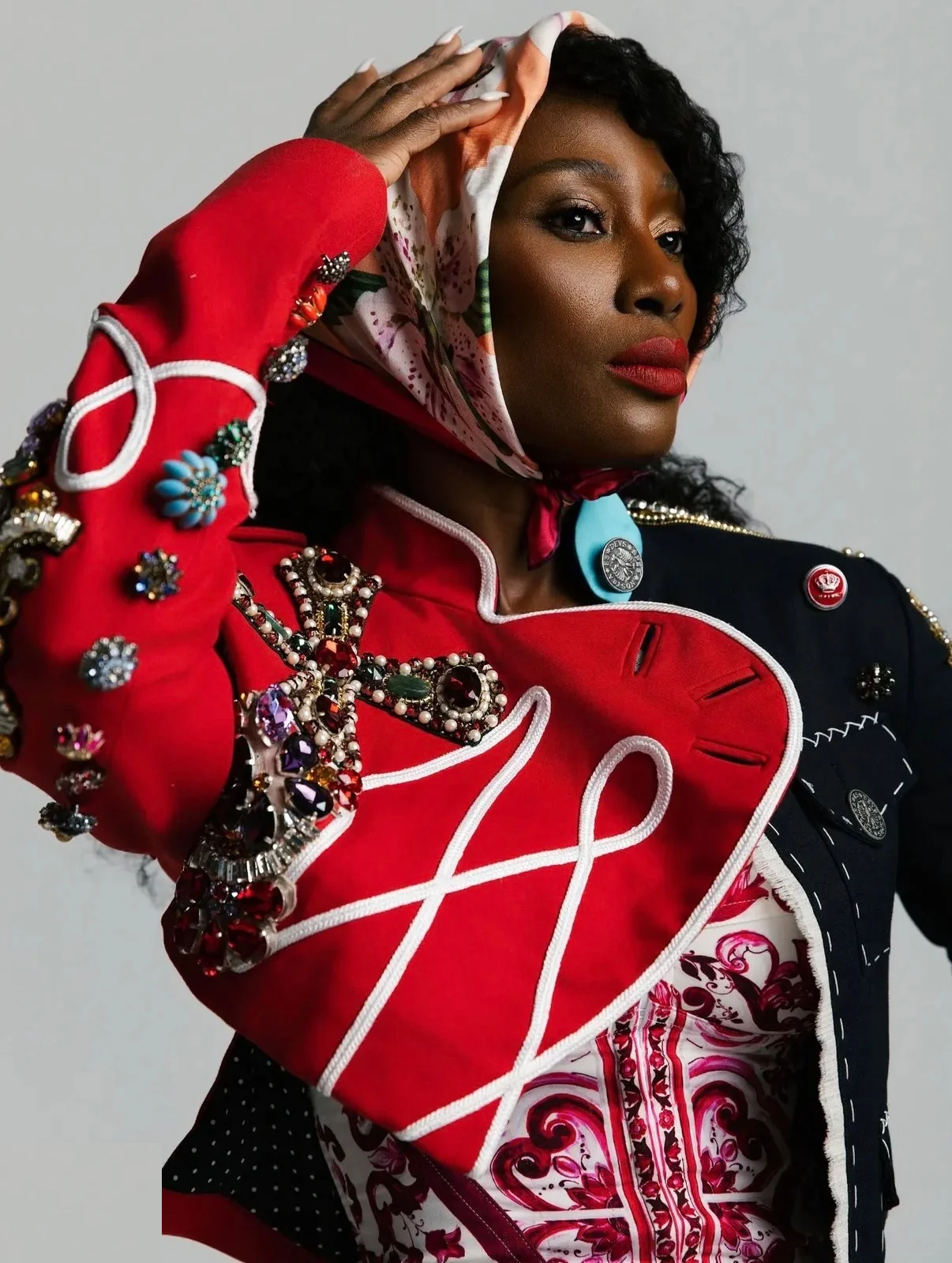 Dede Chancelor in a red-and-navy embellished military jacket with a silk floral headscarf, hand raised to the scarf — studio portrait, grey ground.