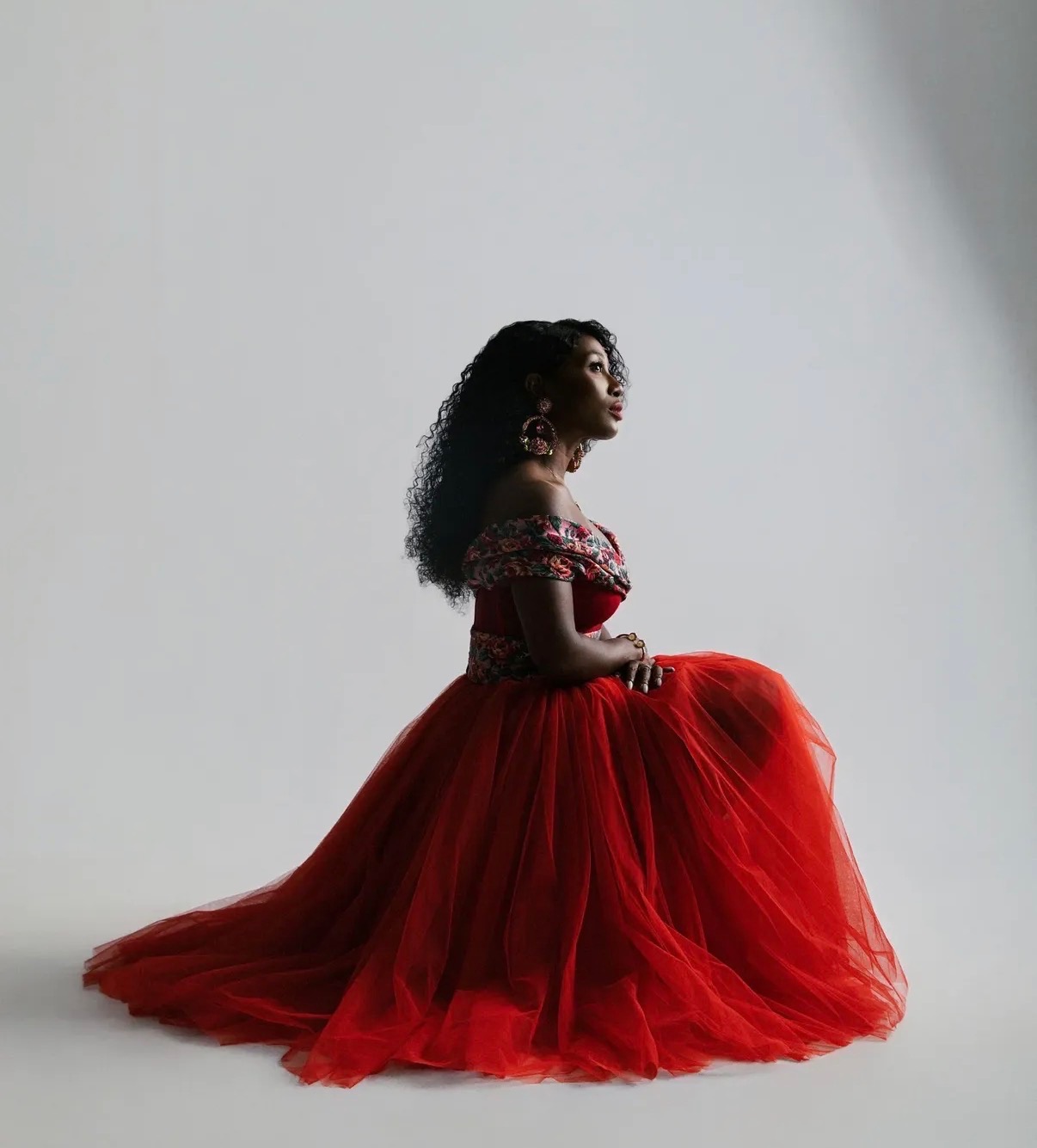 Dede Chancelor seated in a floor-length red tulle gown, studio ground.
